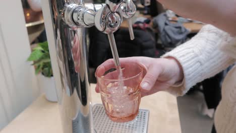 Frau-Gießt-Mineralwasser-In-Ein-Orangefarbenes-Glas-Aus-Einem-Schönen-Chromspender-Im-Innenbereich