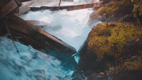 Rushing-Water-Rapids-Running-Through-Log-Chute-and-Crashing-Into-Damaged-Moss-Covered-Log-Chute-Wooden-Beams-While-Spraying-Mist-and-Water-onto-Moss-Covered-Rocks-During-Autumn-4K-ProRes
