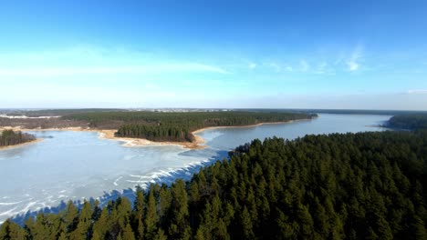 A-beautiful-aerial-backward-shot-of-a-landscape-with-an-enourmous-frozen-lake-and-spruce-forest-during-winter,-Poland