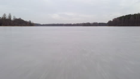 AERIAL:-Flying-Very-Low-and-Close-to-the-Frozen-Icy-Lake-Surface-on-a-Cloudy-Day-with-Forest-Visible-in-the-Horizon