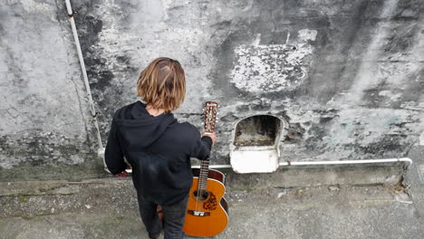 Top-shot-of-a-young-man-wearing-a-black-hoodie-walking-towards-a-classical-guitar,-then-taking-it-with-a-hand-and-holding-it-still,-in-an-abandoned-structure,-on-a-cloudy-day