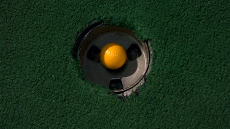 Eine-Nahaufnahme-Eines-Gelben-Minigolfballs-Aus-Der-Vogelperspektive,-Der-In-Das-Golfloch-Fällt-Und-In-Der-Tasche-Auf-Einem-Platz-Abprallt