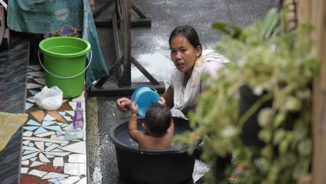 Baby-having-a-bath-with-her-mother-outside-in-wooden-barrel-tub,-female-turns,-smiling-at-camera,-Indonesia