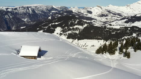 Vista-Aérea-Volando-Hacia-Una-Pequeña-Cabaña-De-Montaña-Cubierta-De-Nieve-En-Los-Alpes-Franceses-En-Invierno