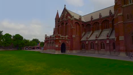 A-low-angle-aerial-view-of-a-beautiful-old-Church-made-with-red-bricks,-Green-grass-and-cars-parked-in-the-parking-of-the-Church
