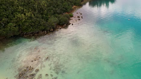 Aerial-view-of-woman-stand-up-paddle-by-shoreline-in-clear-turquoise-water-by-tropical-island