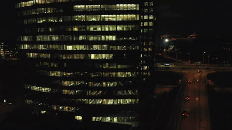 Panning-right-aerial-shot-in-the-dark,-evening-of-an-office-building-with-lights-on-working-over-hours,-and-traffic-with-cars
