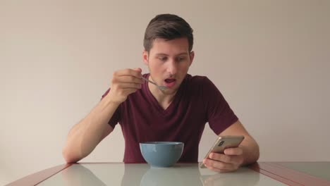 Man-reacting-at-content-on-mobile-phone-while-eating-cereals-sitting-at-glass-table-in-minimalistic-setup-indorrs,-still-medium-shot-eye-level-facing-camera