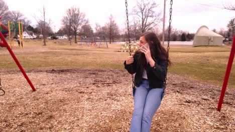 4K-footage-of-a-beautiful,-young,-brunette-college-teenager-talking-on-her-smart-phone-while-sitting-in-a-swing-set-in-a-city-park
