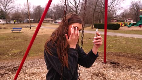 4k-footage-of-a-gorgeous-college-teenager-sitting-on-a-swing-set-using-her-cell-phone-as-a-mirror-to-check-her-makeup-and-hair