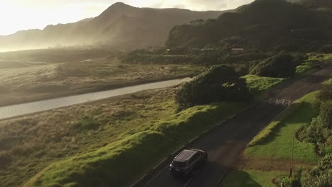 SUV-driving-along-a-thin-road-during-the-golden-hour-at-North-Piha-beach-in-Auckland,-New-Zealand-as-a-misty-lit-horizon-lies-ahead