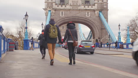 Young-adult-couple-holding-hands-while-crossing-London-Tower-Bridge