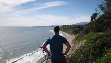 A-man-standing-on-the-edge-of-an-ocean-cliff-on-a-nature-hiking-trail-with-an-epic-beach-view-on-a-sunny-day-in-Santa-Barbara,-California-SLOW-MOTION