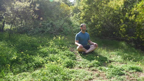A-handsome-man-in-a-green-grassy-forest-meadow-in-nature-on-a-bright-sunny-day-thinking-about-life-in-a-sitting-meditation-pose-to-reduce-stress-and-improve-happiness