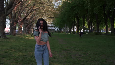 Shot-from-behind-of-an-attractive-and-playful-latina-woman-with-black-wavy-hair-walking-under-the-trees-in-a-park-in-London,-turning-and-looking-at-the-camera,-happy-with-a-beautiful-smile