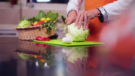 A-slow-mo-side-angle-close-up-view-of-a-lady-chef-cutting-the-cabbage-with-knife-in-the-kitchen,-A-basket-of-veggies-with-tomatoes-and-garlic-on-the-table,-The-chef-wore-and-orange-colored-apron
