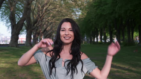 Attractive-and-playful-latina-woman-with-black-wavy-hair-walking-under-the-trees-in-a-park-in-London,-looking-at-the-camera,-spinning-happy-with-a-beautiful-smile
