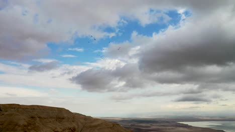 Flock-of-birds-fly-in-rounds-over-Desert-Mountains,-Deadsea-in-the-background,-cloudy-sky,-long-shot