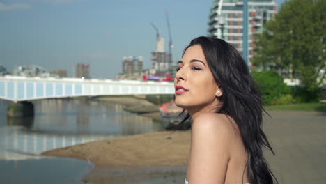 Beautiful-latina-woman-on-holiday-leaning-against-the-railing,-looking-at-the-river-Thames-in-London-and-waving-with-her-hand,-smiling-and-wandering