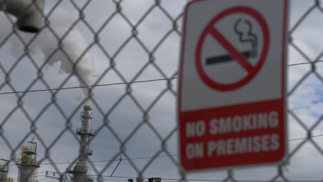 A-refinery-pipe-smoking-and-polluting-in-the-background-with-a-no-smoking-sign-blurred-in-the-foreground-on-a-chain-linked-fence