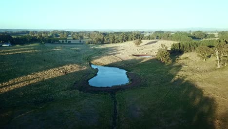 Luftaufnahmen-Abseits-Der-Sonne-Von-Einem-Farmdamm-Und-Runden-Heuballen-Auf-Einem-Feld-In-Der-Nähe-Von-East-Trentham,-Zentral-Victoria,-Australien
