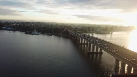 Aerial:-Drone-shot-following-cars-driving-across-a-bridge-towards-the-city-during-morning-rush-hour-as-the-sun-rises-in-Sydney,-Australia
