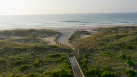 4K-Blick-Auf-Grüne-Dünen-Und-Einen-Hölzernen-Strandzugang,-Der-Bei-Sonnenaufgang-Zu-Einem-Strand-Führt,-An-Dem-Menschen-Spazieren-Gehen