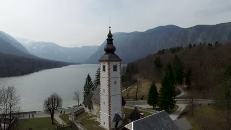 Drone-getting-high-above-of-bridge-and-church-from-the-Bohinj-lake,-Slovenia