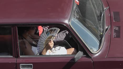 Cuban-women-sitting-on-passenger-seat-of-a-red-vintage-car-while-using-hand-fan-to-cool-the-face-on-a-hot-and-sunny-day-in-the-city-of-Havana