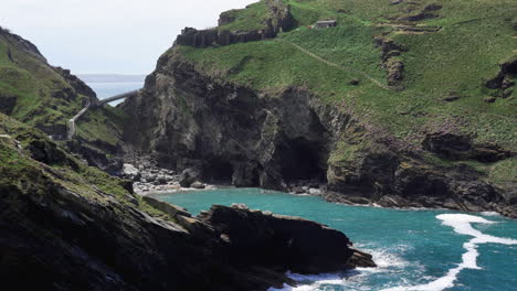 The-ruins-of-popular-tourist-destination-Tintagel-castle-in-Cornwall-on-top-of-a-cliff-with-new-bridge-that-leads-to-the-island-and-the-bay-with-beach-below