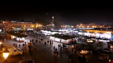 Große-Luftaufnahme-Des-Nachtmarktes-Von-Marrakesch,-Marokko,-Mit-Touristen-Und-Einheimischen,-Die-Die-Souks-Und-Verkaufsstände-Nach-Essen-Und-Souvenirs-Erkunden,-Während-Die-Mondsichel-über-Der-Moschee-Aufgeht