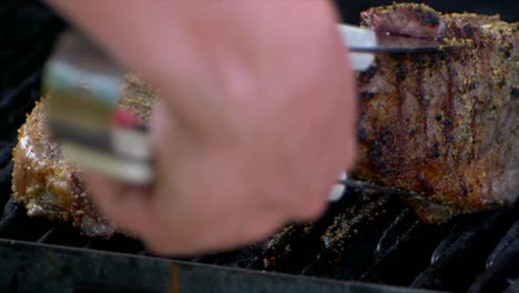 A-pair-of-meat-tongs-turn-a-nearly-cooked-juicy-rib-eye-steak-on-a-grill-and-slides-it-a-bit