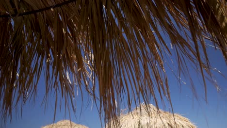 Handheld-Shot-of-Dried-Palm-Leaves-from-a-Tiki-Hut-Umbrella-Moving-By-The-Blowing-Wind-on-a-Sunny-and-Windy-Day-in-Santorini-Greece