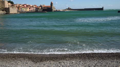 Waves-slowly-hitting-the-beach-in-Collioure-with-the-old-town-in-the-background-on-a-hot-bright-windy-day,-France