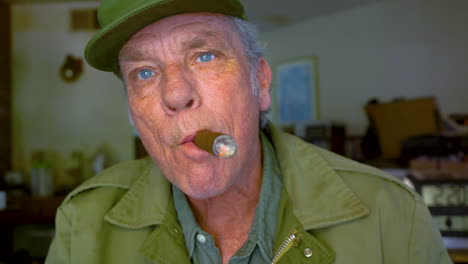 Closeup-of-handsome-mature-male-in-his-60s-smoking-cigar-and-blowing-smoke-directly-at-the-camera
