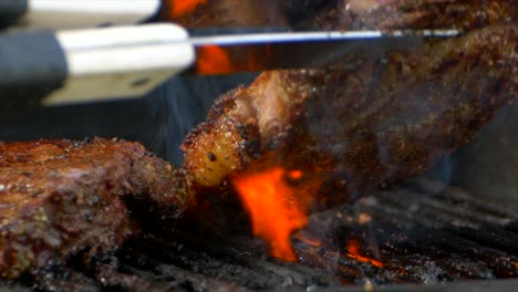 A-pair-of-meat-tongs-turn-a-nearly-cooked-juicy-rib-eye-steak-on-a-grill-as-orange-flames-shoot-up-from-below-in-slow-motion