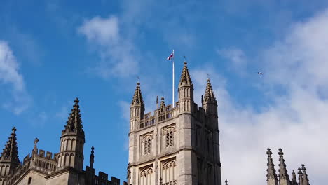 Super-Slow-Motion-Shot-of-Seagull-Flying-Past-Tower-of-Bath-Abbey-in-Somerset,-England-on-Sunny-Summer’s-Day-with-Saint-George’s-Cross-Flapping-in-the-Wind