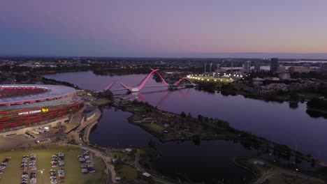Filmische-Luftaufnahme-Eines-Stadions-Am-Swan-River-Mit-Hin--Und-Herbewegung,-Die-Den-Blick-Auf-Die-Skyline-Von-Perth-Bei-Sonnenuntergang-Und-Die-Brücke-Mit-Mäßigem-Verkehr-Freigibt