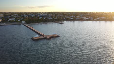 Beautiful-aerial-sunset-over-a-coastal-town-with-long-jetty-for-fisherman,-walkers,-joggers,-family-and-elders-in-Wynnum-Queensland
