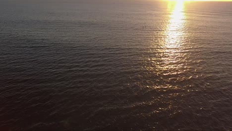 Sunrise-drone-shot-over-water-with-paddle-boarders-passing-through-golden-light-rays