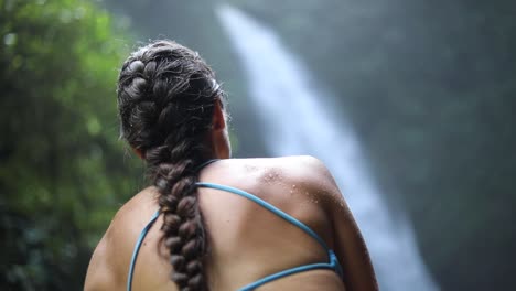Zeitlupenschwenk-Eines-Mädchens-In-Einem-Blauen-Bikini,-Das-Vor-Einem-Sprudelnden-Nungnung-Wasserfall-In-Bali,-Indonesien,-Sitzt