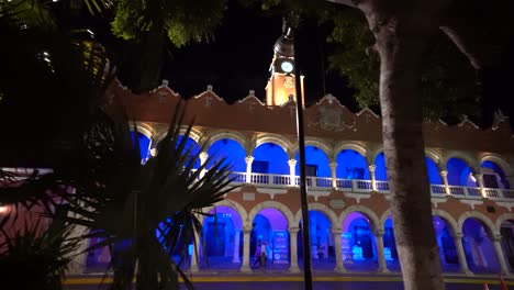 Dolly-shot-to-the-left-of-the-Municipal-building-at-dusk-with-its-blue-lights-next-to-the-plaza-grande-in-Merida,-Mexico