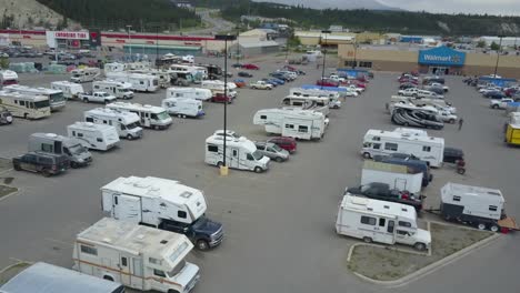 Aerial-footage-of-a-Walmart-parking-lot-in-Whitehorse,-Canada-showing-many-parked-recreational-vehicles-with-cars-moving,-people-walking,-birds-flying,-and-mountains-in-the-background