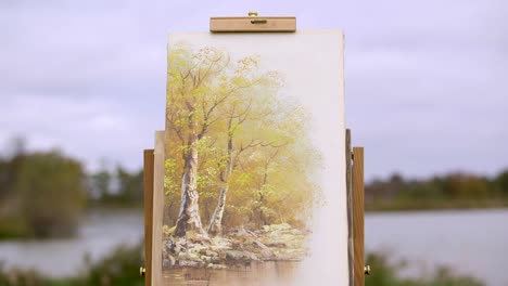 Pintura-De-árboles-Se-Encuentra-Afuera,-Frente-A-Un-Lago-Desenfocado