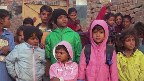 Niños-De-La-Escuela-Del-área-Rural-Paquistaní-De-Pie-Con-Una-Bomba-De-Mano,-En-La-Temporada-De-Invierno,-Usando-Suéteres-Y-Chaquetas,-Ladrillos-Rojos-Y-Una-Vieja-Puerta-En-El-Fondo