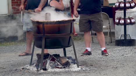 Two-men-standing-as-one-of-them-stirs-steaming-apple-butter-in-a-large-wood-fired-cauldron-outside