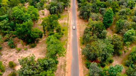 Aerial-view-flying-over-old-patched-two-lane-forest-road-with-car-moving-green-trees-of-dense-woods-growing-both-sides