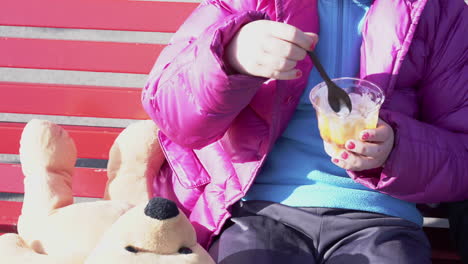 Nahaufnahme-Eines-Kleinen-Mädchens-Mit-Blauem-Pullover,-Nagellack-Und-Teddybär-Neben-Ihr-Mit-Einem-Eisbecher-Im-Freien-4k