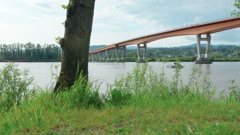 Grasbewachsenes-Flussufer-Mit-Einem-Boot-Und-Einer-Brücke-Im-Hintergrund