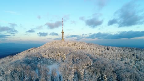 Aerial-view-of-TV-tower-on-mountain-top-surrounded-with-forest-covered-in-snow-at-sunset